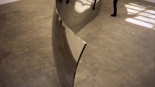 Viewer stands before a large, undulating ribbon of metal on concrete floor. 