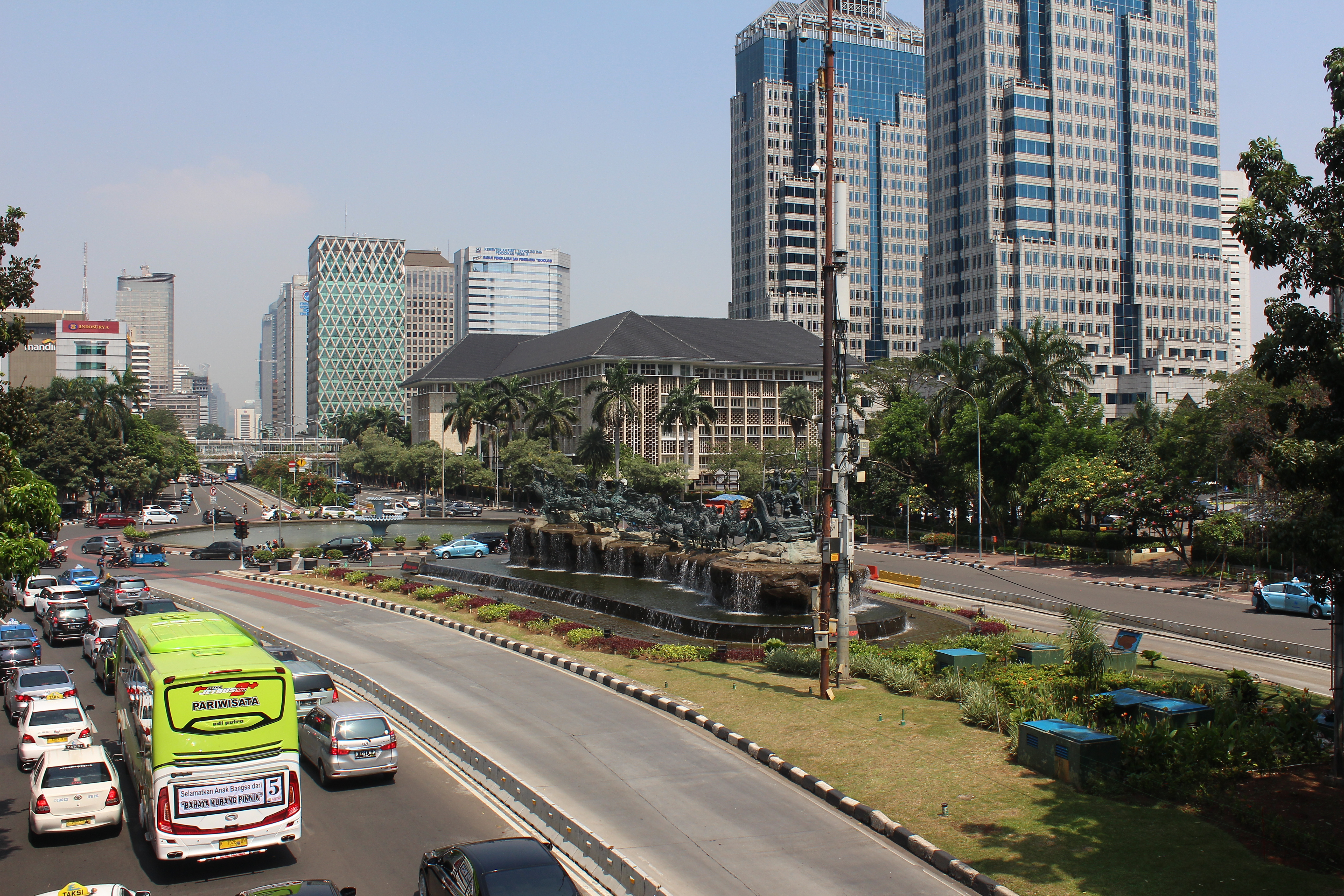 A lawned median dividing a road with cars and a green bus on one side, and tall buildings on the other, with palm trees